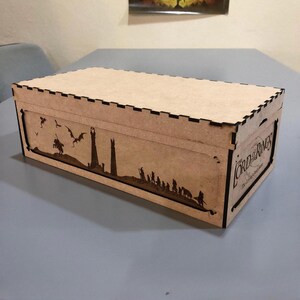 A wooden box with a lid, featuring a silhouette of a castle and a dragon on the front, sitting on a table.