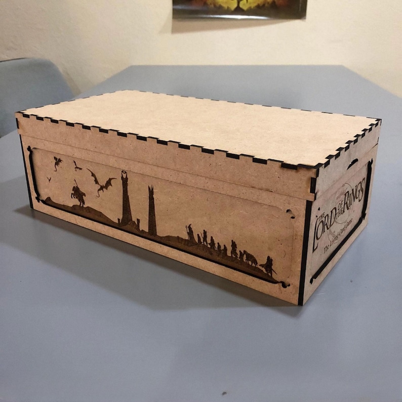 A wooden box with a lid, featuring a silhouette of a castle and a dragon on the front, sitting on a table.