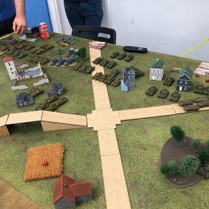 Op de afbeelding: Een tabletop oorlogsgame-opstelling met miniatuur tanks, gebouwen en terrein. Het spelbord heeft een groen oppervlak met wegen, een meer en een brug. Miniatuur tanks zijn in formaties gerangschikt. Een Coca-Cola blikje staat op de tafel.