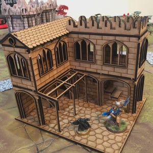 Op de afbeelding: Een lasergesneden houten miniatuurgebouw met een steenachtige gevel, boogvensters en een pannendak. Het model heeft een overdekte veranda en is ontworpen voor tabletop gaming. Inclusief miniatuurfiguren.