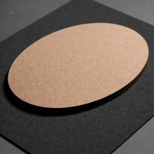 May include: A stack of oval-shaped, light brown wooden cutouts. The edges of the cutouts are visible, showing the layered construction. The cutouts are resting on a dark gray, textured surface.