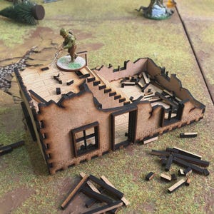 May include: A miniature, laser-cut wooden model of a bombed-out building, with a soldier figurine on the roof. The building has detailed brickwork and exposed interior, with scattered debris. The color palette is primarily brown and tan.
