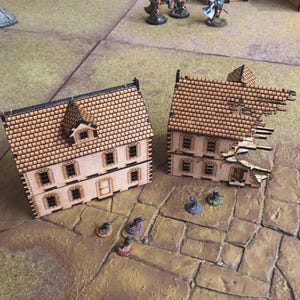 Peut inclure: Deux bâtiments miniatures en bois avec des toits détaillés en forme de briques et des découpes de fenêtres. Un bâtiment est intact, tandis que l'autre a un toit et un mur endommagés. Idéal pour les jeux de table.