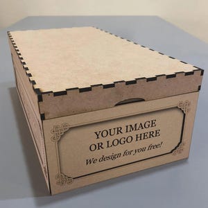 以下が含まれることがあります： 蓋付きの長方形の木製ボックスで、前面パネルに装飾フレームが付いています。ボックスは天然木の色で、縁に黒のアクセントがあります。前面のテキストは「YOUR IMAGE OR LOGO HERE」と「We design for you free!」と書かれています。