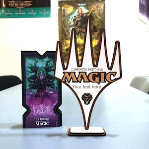 Peut inclure: Trophée en bois Magic the Gathering avec le texte "CAMPEON SEPT-2022 MAGIC" et un espace pour un texte personnalisé. À côté, une carte avec une illustration de personnage fantastique. Le trophée est sur une surface blanche.