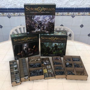 Puede incluir: Un juego de mesa con tres cajas con el título "El Señor de los Anillos: Viajes por la Tierra Media" en español. Las cajas están apiladas, con varias piezas de juego y organizadores visibles. Las piezas del juego son grises y marrones.