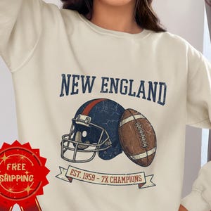 May include: Cream-colored sweatshirt featuring a distressed graphic of a football helmet and football with the text "NEW ENGLAND" and "EST. 1959 - 7X CHAMPIONS". The design is in navy blue, red, and brown.