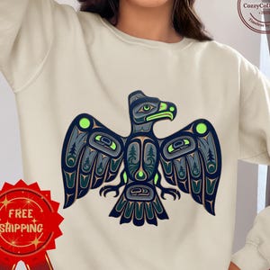 May include: Cream-colored sweatshirt featuring a detailed, stylized eagle design in shades of blue, green, and black. The eagle has outstretched wings and intricate patterns. The sweatshirt also has a "Free Shipping" badge.