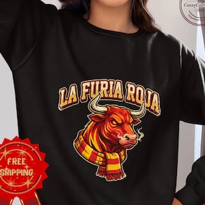 May include: Black sweatshirt featuring a red bull with a yellow and red scarf, and the text "LA FURIA ROJA" in a gold and red font. The bull has a fierce expression. The sweatshirt has a round neck.