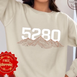 May include: Cream-colored sweatshirt featuring the number "5280" in large white letters above a mountain range graphic. The graphic is in shades of brown and red. The sweatshirt has a crew neck and long sleeves.