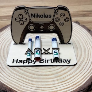 Könnte beinhalten: Ein hölzerner Tortenaufsatz zum Geburtstag mit einem Videospiel-Controller-Design, graviert mit dem Namen "Nikolas". Der Aufsatz zeigt die Zahlen "50" und die Worte "Happy Birthday". Er steht auf einer Holzscheibe.