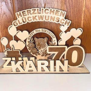 Puede incluir: Una decoración de cumpleaños de madera con las palabras "Herzlichen Glückwunsch" y "Happy 70th Birthday" junto con el nombre "Karin" y el número "70". La decoración presenta un leopardo y corazones.
