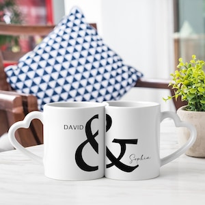 May include: Two white ceramic mugs with heart-shaped handles. One mug has the name "David" printed on it, and the other has the name "Sophia". A large black ampersand symbol is between the names. The mugs are on a white surface.