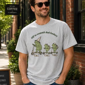 May include: A light gray t-shirt with the phrase "Off to commit dad foolery" and a graphic of three frogs on scooters. The frogs are labeled "Dad", "Emily", and "James". The person is wearing sunglasses and blue jeans.