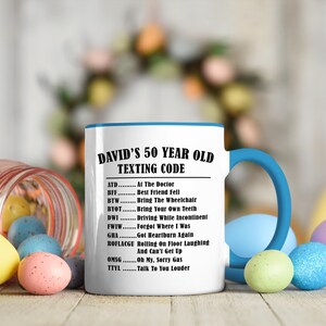 May include: A white ceramic mug with a blue handle and rim, featuring the text "DAVID'S 50 YEAR OLD TEXTING CODE" and a list of acronyms with their meanings. The mug is surrounded by colorful Easter eggs, suggesting a gift theme.