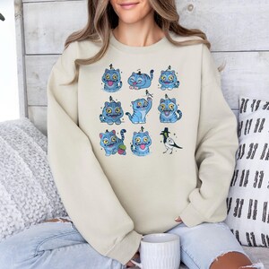 May include: A cream-colored sweatshirt featuring a repeating pattern of cartoon blue cats with yellow eyes and a black bird. The cats have various expressions and poses. The sweatshirt is a casual style.