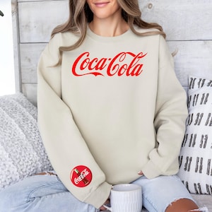 May include: A beige sweatshirt with the Coca-Cola logo in red script across the chest. A smaller Coca-Cola logo is on the left sleeve. The sweatshirt is a crew neck style.