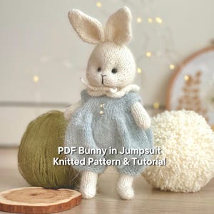 Könnte beinhalten: Eine gestrickte Hasenpuppe mit einem hellblauen Overall mit Rüschenkragen und zwei kleinen Knöpfen. Der Hase hat lange Ohren und ein schwarz gesticktes Gesicht. Der Text "PDF Bunny in Jumpsuit Knitted Pattern & Tutorial" ist sichtbar.