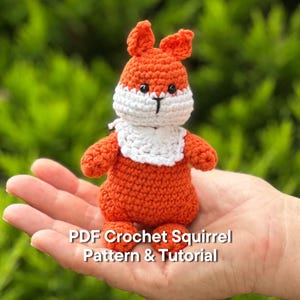 Puede incluir: Un juguete de ardilla de ganchillo en tonos naranja, blanco y negro. La ardilla tiene un babero blanco y una nariz y ojos negros. El texto "PDF Crochet Squirrel Pattern & Tutorial" se muestra en la parte inferior.