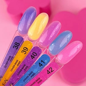 Puede incluir: Una exhibición de seis muestras de esmalte de uñas en varios colores, incluyendo morado, amarillo, rosa y azul. Cada muestra está montada en un palito de plástico transparente con un número e información del producto. El fondo es rosa brillante.