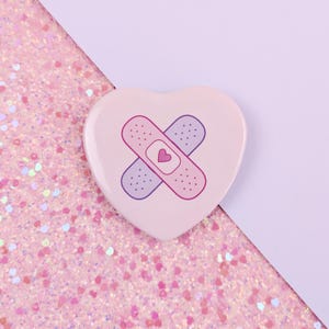 May include: A heart-shaped pin with a light pink base color. The center of the pin features a graphic illustration of two adhesive bandages crossed over each other in light purple and pink, with a small pink heart icon in the center of the bandages.
