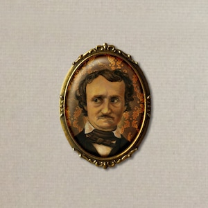 Könnte beinhalten: Eine ovale Brosche aus Bronze mit einem Kamee von Edgar Allan Poe. Das Kamee zeigt ein Porträt von Poe mit einem dunkelbraunen Schnurrbart und einem dunkelbraunen Bart. Der Hintergrund des Kamees ist ein braunes und orangefarbenes Blumenmuster.