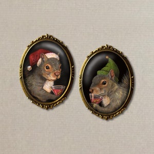 May include: Two oval-shaped brooches with a black background and gold trim. Each brooch features a gray squirrel wearing a festive hat and holding a red cocktail glass. One squirrel wears a red Santa hat, the other a green elf hat.