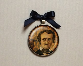 Puede incluir: Un colgante redondo dorado con un retrato de Edgar Allan Poe y un cuervo. El colgante está unido a una cinta negra.