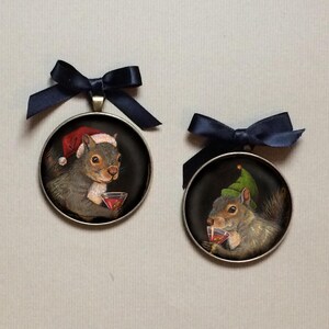 May include: Two round ornaments with black ribbon hangers. Each ornament features a painted image of a squirrel wearing a festive hat and holding a glass of red liquid. One squirrel wears a Santa hat, the other a green elf hat.