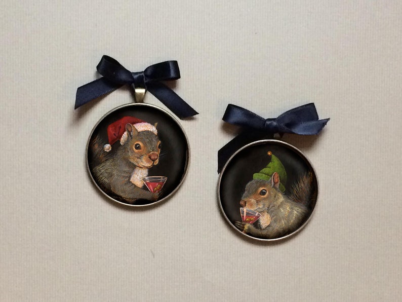May include: Two round ornaments with black ribbon hangers. Each ornament features a painted image of a squirrel wearing a festive hat and holding a glass of red liquid. One squirrel wears a Santa hat, the other a green elf hat.