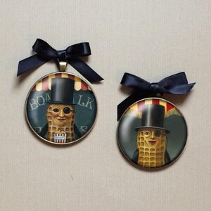 May include: Two round pendants featuring the Mr. Peanut character, each with a black top hat and monocle. The pendants have a dark blue ribbon bow at the top. The background of the pendants reads "Boardwalk Peanuts".