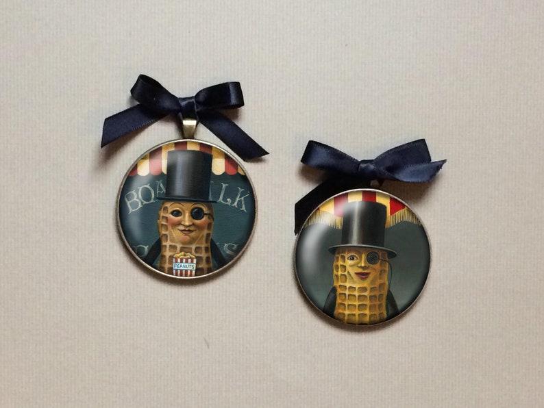 May include: Two round pendants featuring the Mr. Peanut character, each with a black top hat and monocle. The pendants have a dark blue ribbon bow at the top. The background of the pendants reads "Boardwalk Peanuts".