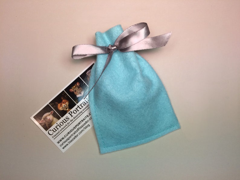Peut inclure: Un sac en feutre bleu clair avec un ruban argent&eacute; nou&eacute; autour du haut. Le sac est pos&eacute; sur une surface blanche. Une carte de visite pour "Curious Portraits" est visible en arri&egrave;re-plan.