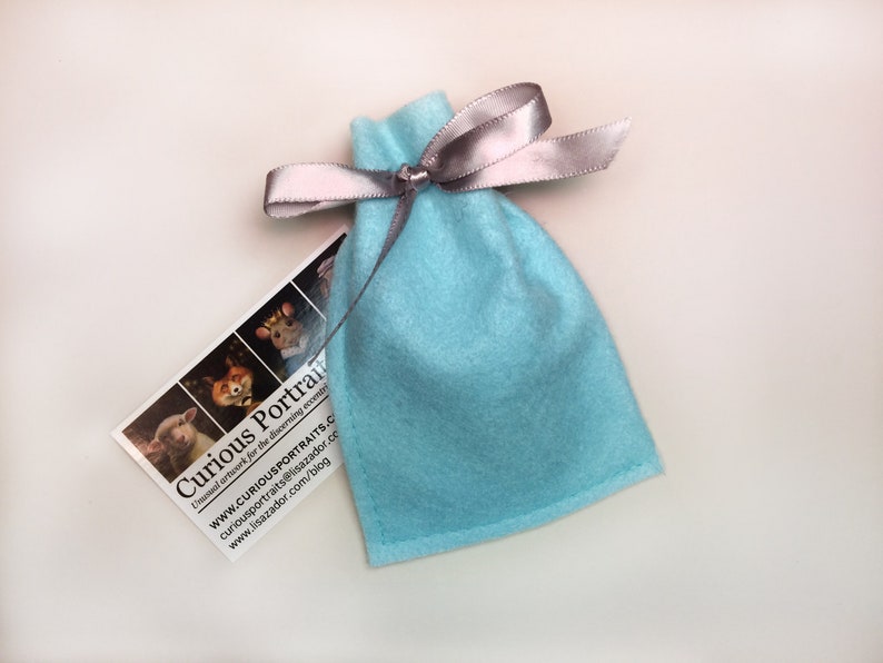 May include: A light blue felt bag with a silver ribbon tied around the top. The bag is sitting on a white surface. A business card with the text "Curious Portraits" is visible in the lower left corner of the image.