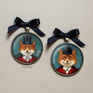 May include: Two round, gold-toned ornaments with black ribbon hangers. Each ornament features a fox wearing a different hat. One fox wears a top hat and the other wears a riding helmet. Both foxes are wearing a red jacket and a white bow tie. The background is a blue-green color.