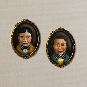 May include: Two vintage-style portrait brooches featuring painted images of wooden dolls with painted faces and bow ties. The brooches are in gold-toned oval frames with ornate details.