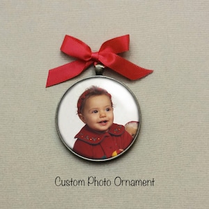 Peut inclure: Une d&eacute;coration ronde en argent avec un ruban rouge. La d&eacute;coration pr&eacute;sente une photo d'une petite fille portant un manteau rouge et un bandeau rouge. Le texte "Custom Photo Ornament" est imprim&eacute; sous la d&eacute;coration.