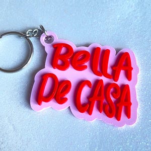 Può includere: Un portachiavi rosa con le parole "Bella De Casa" in corsivo rosso. Il portachiavi ha un anello e un fermaglio argentati per attaccarlo alle chiavi o a una borsa. Lo sfondo è una superficie chiara e glitterata.