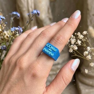 Könnte beinhalten: Ein leuchtend blauer Ring mit dem weißen Schriftzug "CORE de MAMMA". Der rechteckige Ring wird an einem Finger getragen. Der Hintergrund ist unscharf, mit Blumen.