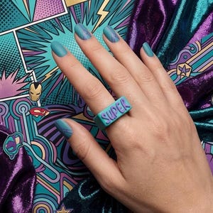 Può includere: Un anello turchese con la scritta "SUPER" in lettere viola è indossato su una mano con smalto turchese. Le unghie hanno un disegno viola. Lo sfondo presenta un'arte in stile fumetto con immagini di supereroi e un tessuto viola lucido.