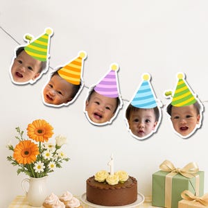 May include: A birthday banner with smiling baby faces wearing party hats in various colours. The banner hangs above a table with a chocolate cake, flowers, cupcakes, and a wrapped gift.