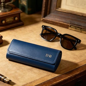 Peut inclure: Un étui à lunettes rectangulaire en cuir bleu marine avec une fermeture à rabat et des coutures blanches. Les initiales DB sont gravées en doré sur le devant. À côté de l'étui se trouve une paire de lunettes de soleil à monture noire.