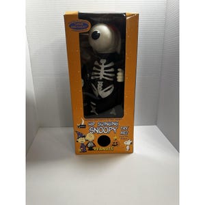 May include: A Halloween-themed Snoopy toy in a skeleton costume, packaged in an orange box. The toy has a large, white eyeball and a black skeleton suit. The box features the text "Hip Swinging Snoopy" and "Peanuts."