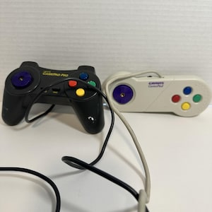 May include: Two vintage game controllers. One is black with the text "GAMEPAD PRO" and colorful buttons. The other is off-white with the text "GRAVIS GamePad" and colorful buttons. Both controllers have attached cords.