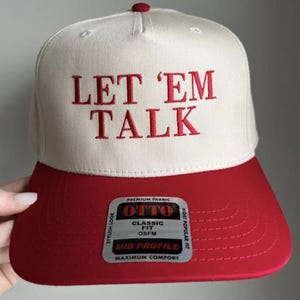 May include: A cream and red baseball cap with the embroidered text "LET 'EM TALK" in red. The cap has a red brim and a small red button on top. The label reads "OTTO CLASSIC FIT OSFM MID PROFILE MAXIMUM COMFORT".