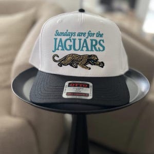 May include: A white baseball cap with a black brim featuring the embroidered teal text "Sundays are for the JAGUARS" above a graphic of a prowling jaguar in gold, black, and teal. An Otto brand sticker is centered on the flat black brim.