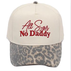 May include: A cream-coloured baseball cap with the embroidered text "All Sugar No Daddy" in red. The brim features a leopard print pattern in shades of grey and tan. The cap has a grey button on top.