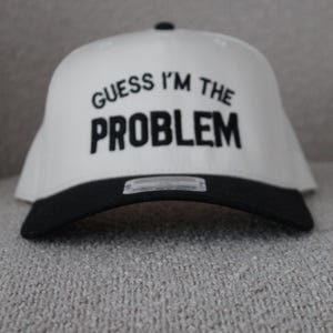 May include: A white baseball cap with a black peak, featuring the text "GUESS I'M THE PROBLEM" in black. The cap is displayed on a textured grey surface.