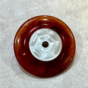 May include: A circular button featuring a wide, polished amber-colored outer ring with a mottled texture and a smaller, centered white disc. The white center has a raised geometric pattern with a dark, circular indentation at the very middle.