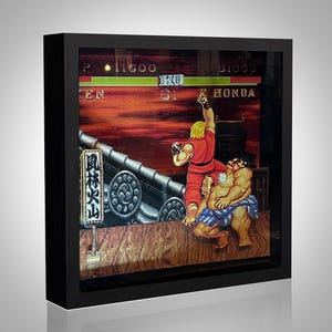 May include: Framed artwork featuring a scene from the video game Street Fighter II. The artwork depicts a fight between Ken and E. Honda, with a red and orange sunset background. The frame is black.